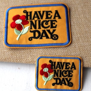Peut inclure: Deux patchs brodés à repasser avec un fond jaune, une bordure bleue et une fleur rouge avec une tige verte. Le texte sur les patchs est "Have a Nice Day".
