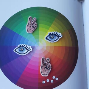 May include: Embroidered patches featuring a peace sign and an eye design. The patches are in shades of brown and blue, with white outlines. The patches are arranged on a colorful gradient background, with a color wheel design.