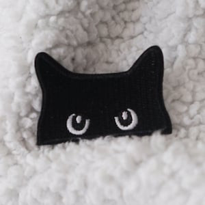 Könnte beinhalten: Schwarzer Filz-Katzenkopf-Patch mit weißen Augen. Der Patch befindet sich auf einer weißen, flauschigen Oberfläche.