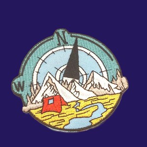 May include: A circular embroidered patch featuring a compass design with the letters N and W. The graphic depicts a tall black mountain peak, white snow-capped mountains, a small red tent, and a winding blue river set against a yellow landscape.