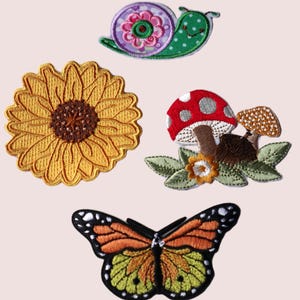 May include: Four embroidered iron-on patches featuring a sunflower, a snail, a mushroom, and a butterfly. The sunflower is yellow with a brown centre. The snail is green with pink and white accents. The mushroom is red with white dots and a brown stem. The butterfly is orange, yellow, and black.