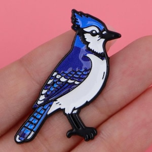 May include: A blue jay bird pin with a black outline. The bird has blue, white, and black details. The pin is a detailed, colourful representation of the bird.
