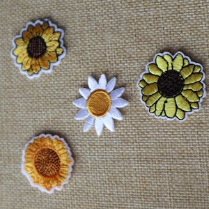 Könnte beinhalten: Fünf gestickte Bügelbilder mit gelben Sonnenblumen und einer weißen Margerite. Die Patches befinden sich auf einem braunen Jute-Hintergrund.