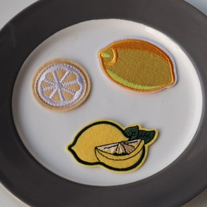 May include: Three embroidered lemon patches on a white plate. One patch is a whole lemon, another is a lemon slice, and the third is a lemon wedge.