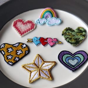 May include: A selection of embroidered patches in heart, star, and rainbow shapes. The patches display diverse colours and patterns, including a pink heart with white stitching, a yellow heart with white flowers, and a star with gold and white detailing.
