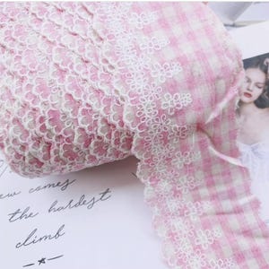 May include: A roll of pink and white gingham fabric with a delicate white floral lace trim. The lace features a repeating pattern of small flowers. The fabric is soft and appears to be cotton.