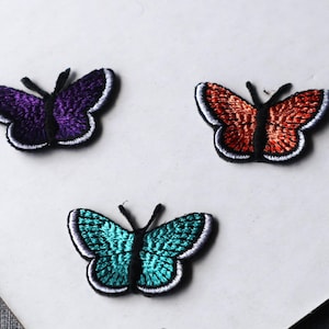 May include: Three embroidered butterfly patches featuring a purple, an orange, and a teal butterfly. Each butterfly has a black central body, black antennae, and a thin white border outlining the wings, with detailed thread texture throughout.
