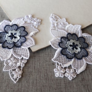 May include: Two white lace floral appliques with a layered design. Each applique features a central flower in shades of blue and gray, surrounded by white lace petals and leaves. The appliques are on a gray surface.