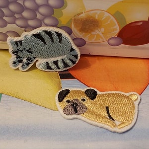 May include: Two embroidered iron-on patches, one of a sleeping cat and one of a sleeping pug. The cat is light blue with black stripes and the pug is light brown with black ears and nose.