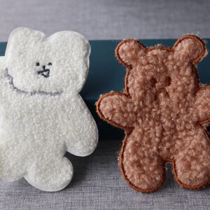 May include: Two plush bear-shaped patches. One is white with a stitched smile and eyes. The other is brown with a stitched outline. These patches could be used for clothing or crafts.