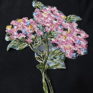 May include: A pink and white embroidered flower applique with green leaves and a blue stem. The applique is on a black fabric background.
