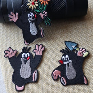 May include: Three embroidered patches featuring a cartoon mole character. The first patch shows the mole holding a bouquet of flowers. The second patch shows the mole with its arms raised. The third patch shows the mole holding a shovel.