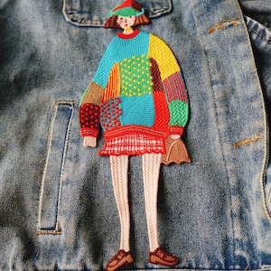 May include: A colorful embroidered patch of a stylized figure wearing a patchwork sweater, red skirt, and brown shoes. The figure is holding a small purse. The patch is affixed to a denim jacket.