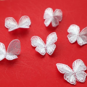 May include: Six white embroidered butterfly appliques with detailed wing patterns are arranged on a red surface. The butterflies vary in size and wing design, showcasing delicate threadwork. These appliques are suitable for craft projects.