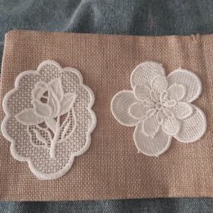 May include: Two white lace flower appliques on a brown hessian background. The appliques are delicate and intricate, with a floral design.