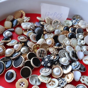 May include: A collection of assorted buttons in a white bowl on a red surface. The buttons vary in size, shape, and color, including black, white, gold, and pearl. Some buttons have decorative embellishments. A small piece of paper reads "120 PCS".