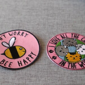 Peut inclure: Deux écussons ronds brodés. L'un, rose, représente une abeille de dessin animé avec le texte « DON'T WORRY BEE HAPPY ». L'autre, rose, dit « I LOVE ALL THE CATS IN THE WORLD » avec des illustrations de chats.