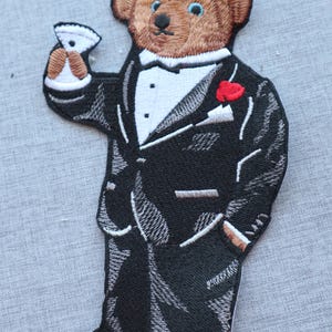 Peut inclure: Écusson brodé d'un ours en peluche brun portant un smoking noir, une chemise blanche et un nœud papillon. L'ours tient un verre à martini et porte une rose rouge. Le patch est cousu avec du fil blanc.