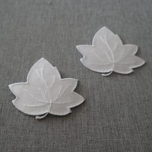 May include: Two white leaf-shaped fabric appliqués featuring delicate embroidered vein details. Each piece is crafted from a sheer, lightweight material with a clean-cut edge, showcasing a symmetrical botanical design with a pointed stem base.