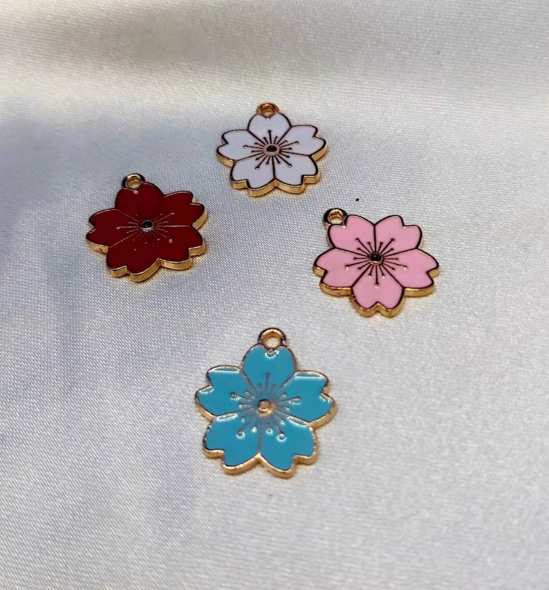 May include: Four gold-colored metal charms shaped like cherry blossoms. The charms are painted in different colors: white, red, pink, and blue.