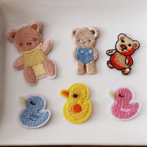 May include: Six embroidered iron-on patches featuring a variety of cute animal designs. The patches include a blue duck, a pink duck, a yellow duck, a red bear, a brown bear wearing a blue swimsuit, and a brown bear wearing a yellow swimsuit.
