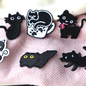 May include: A selection of black cat patches in various poses and designs. One cat is in a coffee cup, another has a pink bow, and others are in different poses. The patches are on a pink fabric background.