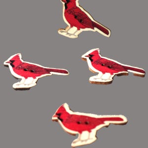 May include: Four wooden cardinal bird cutouts. Each bird is primarily red with black detailing and a white base. The birds are in various poses, with a grey background.