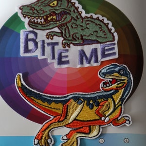 May include: Two embroidered iron-on patches featuring dinosaurs. The first patch is a green dinosaur with the text "Bite Me" in purple. The second patch is a yellow, orange, and blue dinosaur.