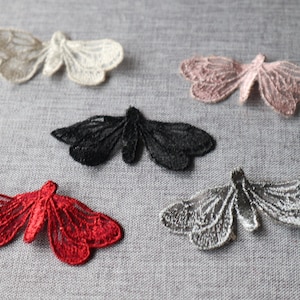 May include: Five embroidered butterfly appliques in various colors: off-white, pink, black, red, and silver. The delicate lace-like wings are detailed and spread, creating a decorative element for crafts and embellishments.