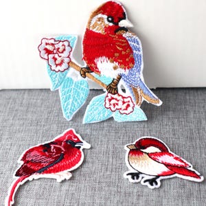 May include: Three embroidered iron-on patches featuring red and blue birds. One bird is perched on a branch with blue leaves and pink flowers. The other two birds are on white backgrounds.