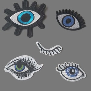 May include: Five embroidered eye patches in different designs. The patches feature blue, green, and white eye details with black outlines. One patch has a starburst shape around the eye.