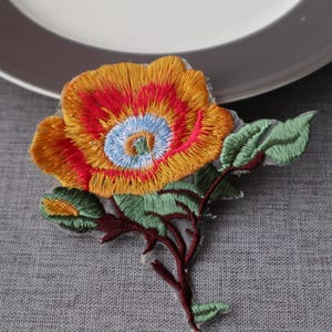 May include: Embroidered floral patch showing an orange, red, and yellow flower with a blue centre. Green leaves and a brown stem complete the design. The patch is likely for decorative purposes.