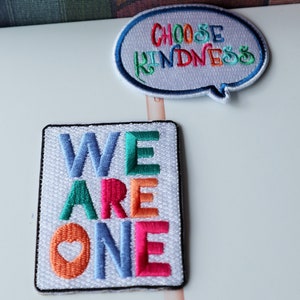May include: Two embroidered patches. One is a light gray speech bubble shape with the text CHOOSE KINDNESS in multicolored thread. The other is a white rectangular patch with the text WE ARE ONE in blue, green, orange, and pink thread.
