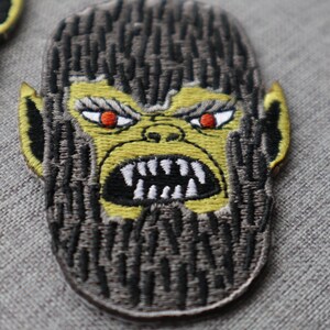 May include: Embroidered patch depicting a stylized ape face with an angry expression. The ape has a yellow face, red eyes, and a black open mouth with white teeth. The patch has a brown and black fur-like texture.