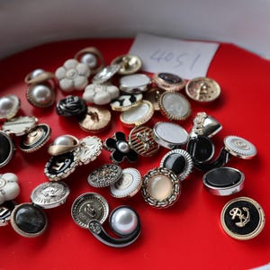 May include: A selection of decorative buttons in diverse designs and colours, including pearl, floral, and metallic details. The buttons are arranged on a red surface. A small white label displays the number "4051".
