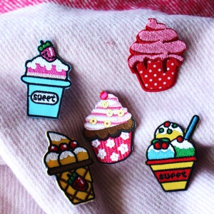 Peut inclure: Cinq patchs brodés colorés à repasser représentant différents desserts sucrés : un cupcake avec un glaçage rose et des pois rouges, un cupcake avec un glaçage brun et des fleurs roses, un sundae avec de la glace bleue et verte, un cornet de gaufres avec trois boules de glace et un gobelet à glace bleu et blanc avec une fraise sur le dessus. Chaque patch est brodé du mot « sweet ».
