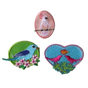 May include: Three embroidered iron-on patches featuring birds. One patch is pink and oval shaped with a white cockatoo. Another patch is green and round with a blue bird on a branch with pink flowers. The last patch is a blue heart shape with two pink birds perched on a branch with green leaves.