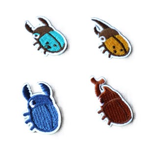 Könnte beinhalten: Vier gestickte Bügelbilder mit verschiedenen farbigen Käfern. Die Käfer sind braun, blau, türkis und gelb.