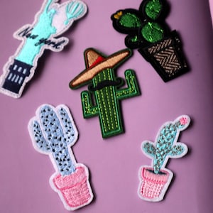 May include: Five embroidered iron-on patches featuring different designs. The patches include a cactus wearing a sombrero, a cactus with blue and white accents, a cactus with green accents, a cactus with pink accents, and the Statue of Liberty with the text "New York" on it.