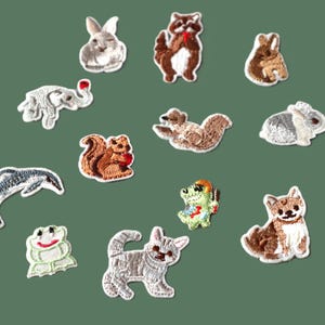 May include: A selection of embroidered animal patches, featuring a rabbit, raccoon, elephant, squirrel, dolphin, frog, cat, and dog. The patches are intricately stitched and colourful, set against a green backdrop.