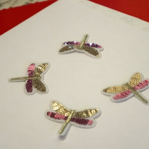 Könnte beinhalten: Vier goldene und rosafarbene Pailletten-Libellen-Patches. Die Libellen haben einen weißen Umriss und sind in einem Kreis auf weißem Hintergrund angeordnet.