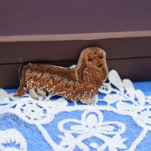 May include: Embroidered patch of a brown dachshund dog with detailed stitching. The dog has long fur and is in a standing position. The patch is on a blue surface with white lace trim.