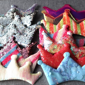 May include: Assortment of colourful sequined crowns in various styles and designs. The crowns are made of fabric and sequins and feature different colours, including pink, red, blue, green, and silver.