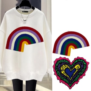 May include: White sweatshirt with a rainbow embroidered on the front. The rainbow is made of purple, blue, green, red, and yellow fabric. There is a heart-shaped patch with a rainbow design on the bottom right corner of the image.