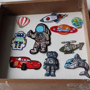 May include: A selection of embroidered patches with diverse designs. Includes a red race car, a space shuttle, a helicopter, robots, an astronaut, a dolphin, and hot air balloons. The patches are displayed in a wooden tray.