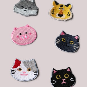 Könnte beinhalten: Sechs gestickte Bügelbilder mit Tiergesichtern. Die Patches sind in verschiedenen Farben, darunter rosa, grau, schwarz, weiß und gelb. Die Tiere umfassen einen Wolf, einen Tiger, ein Schwein, eine Katze und zwei schwarze Katzen.