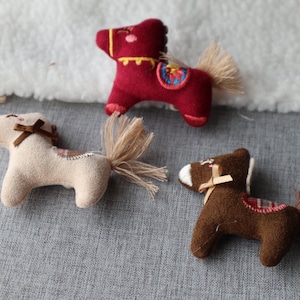 May include: Three small, handmade toy horses in shades of red, tan, and brown. Each horse has a decorative tail and a small bow. The horses are placed on a gray surface.