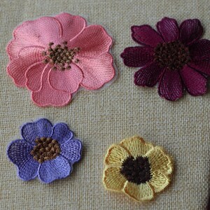 May include: Four embroidered fabric flower patches in pink, purple, burgundy, and yellow. The patches are on a beige fabric background.