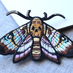 May include: Embroidered moth patch with a skull-like design on the head. The patch features a detailed design with black, brown, gold, blue, and purple colours. The moth's wings are spread, showcasing intricate patterns. This decorative patch can be applied to clothing or accessories.