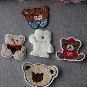 May include: Five embroidered teddy bear patches in various designs and colours. The bears are brown, white, and tan, with red and blue accents. Each patch has a white border. Ideal for clothing or accessories.